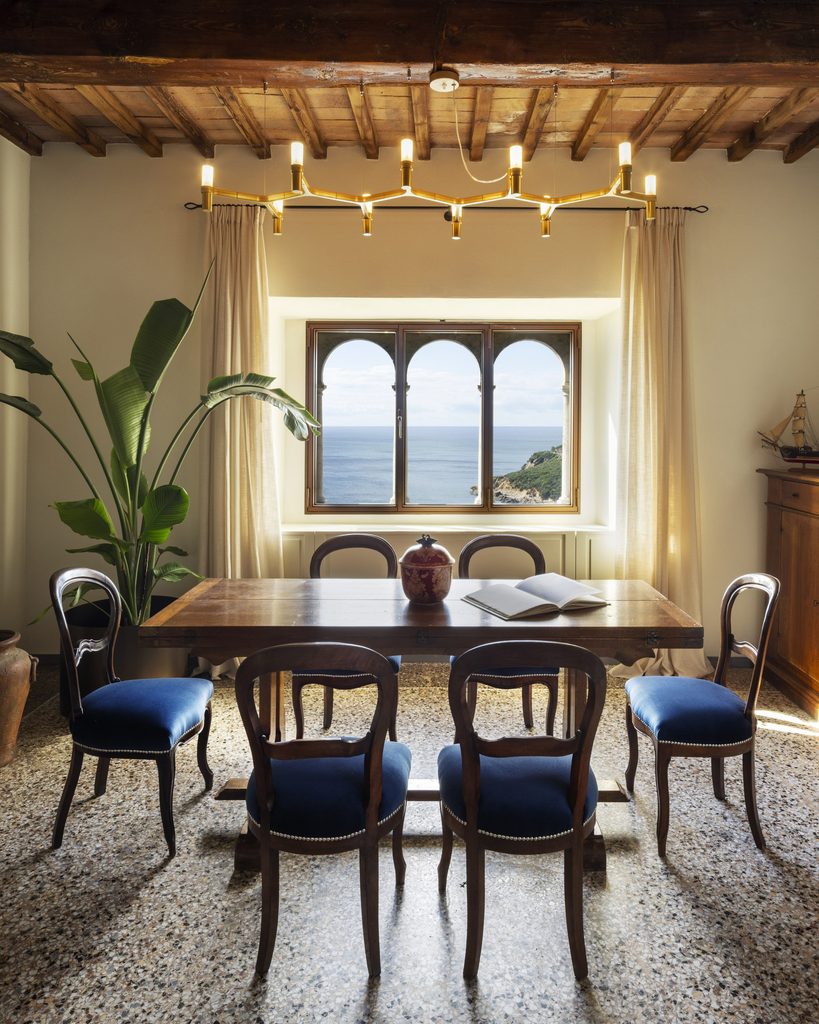 Sala da pranzo di Castel Sonnino con soffitto a travi in legno, sedie in velluto blu, pavimento in seminato veneziano e trifora affacciata sul mare.