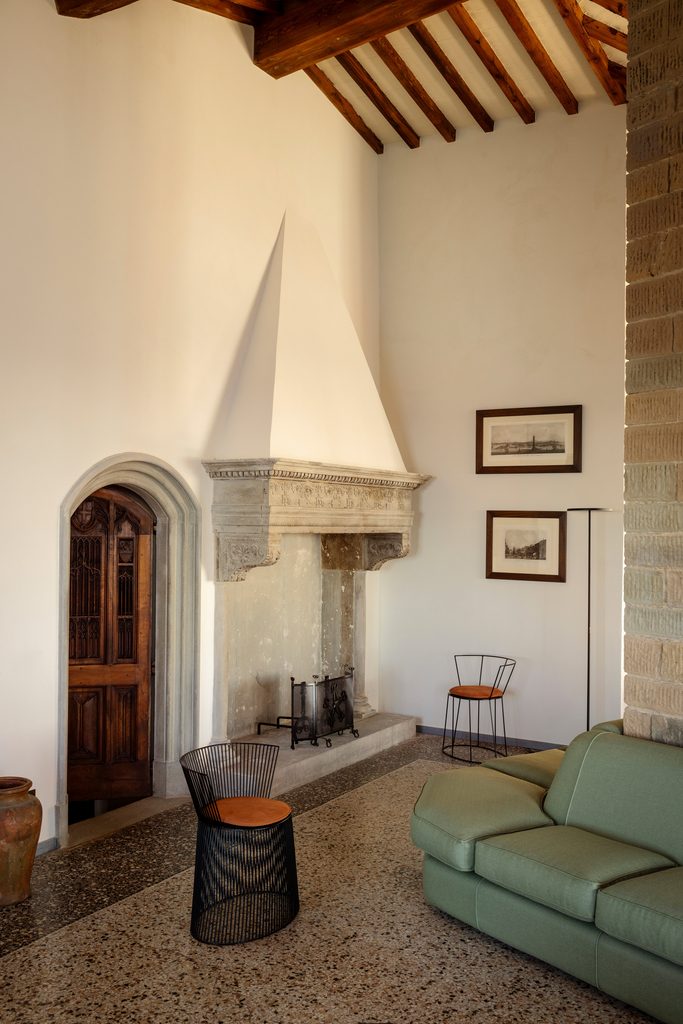 Salotto con grande camino in pietra, porta ad arco in legno, divano verde e pavimento in seminato veneziano.