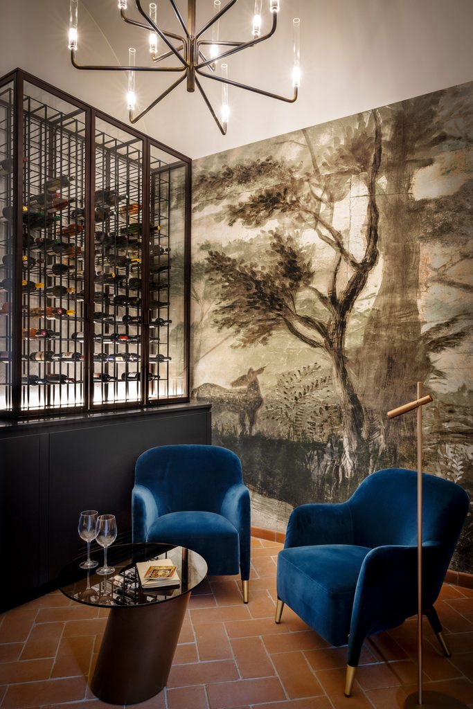 Angolo degustazione con cantina a vista in metallo e vetro, poltrone in velluto blu e parete decorata a paesaggio
