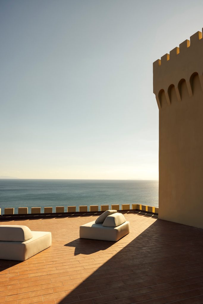 Terrazza in cotto ai piedi della torre merlata, con sedute chiare e vista sul mare al tramonto