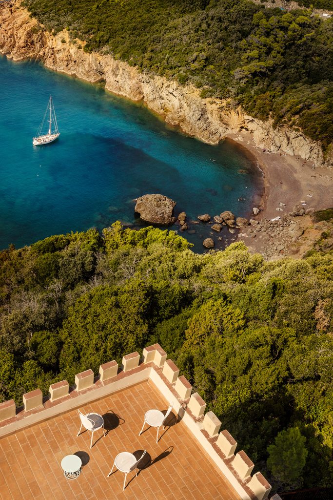 Cala con spiaggia e barca a vela vista dall’alto, con la terrazza merlata di Castel Sonnino in primo piano
