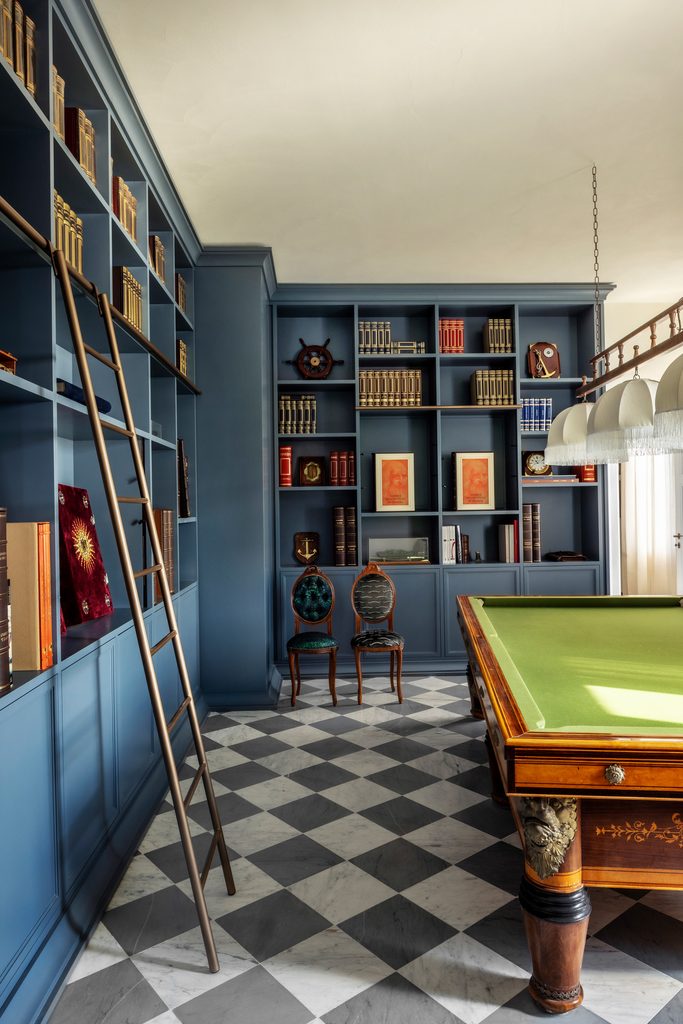 Biblioteca con biliardo d'epoca: libreria su misura blu con scala, pavimento a scacchi in marmo bianco e grigio