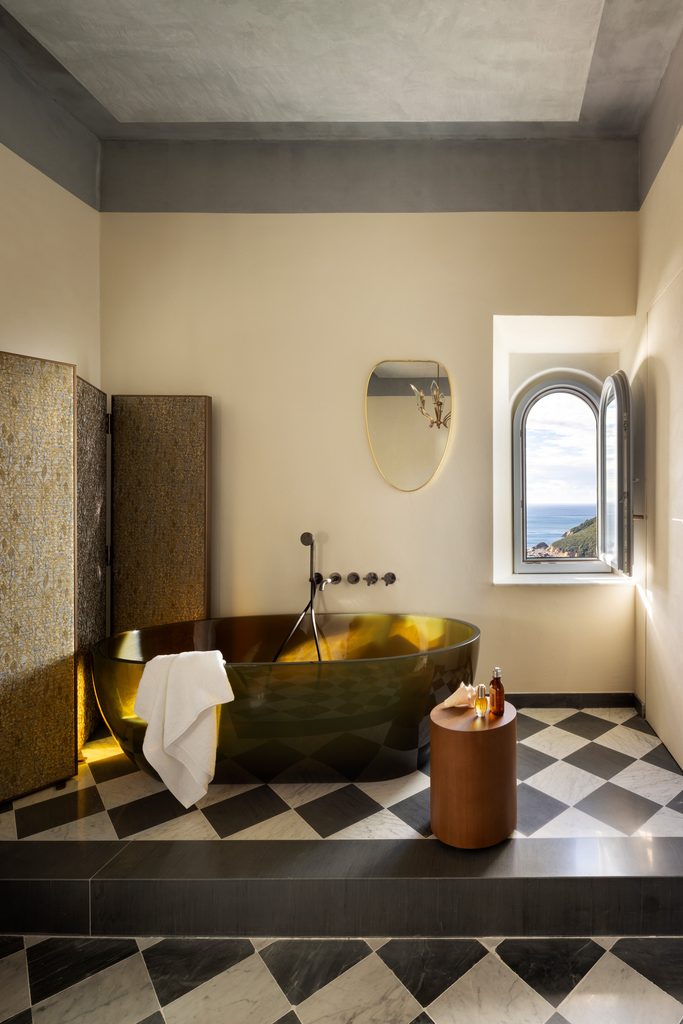 Bagno con vasca freestanding color ambra su pedana, paravento decorato, pavimento a scacchi e finestra ad arco sul mare