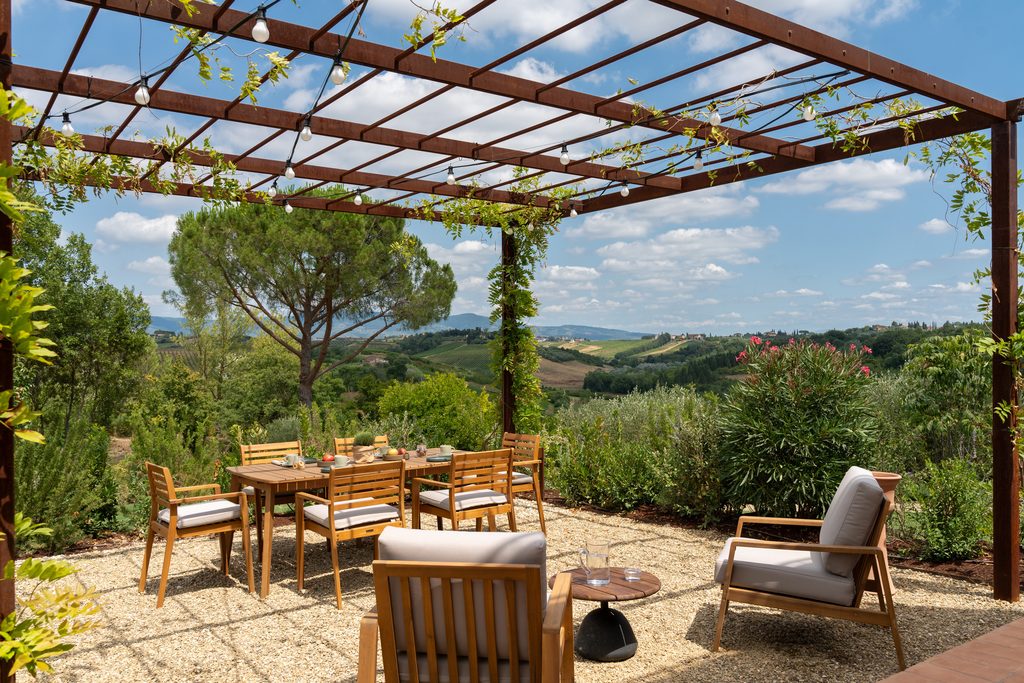 Pergola con rampicanti e tavolo da pranzo su ghiaia, aperta sul panorama delle colline.