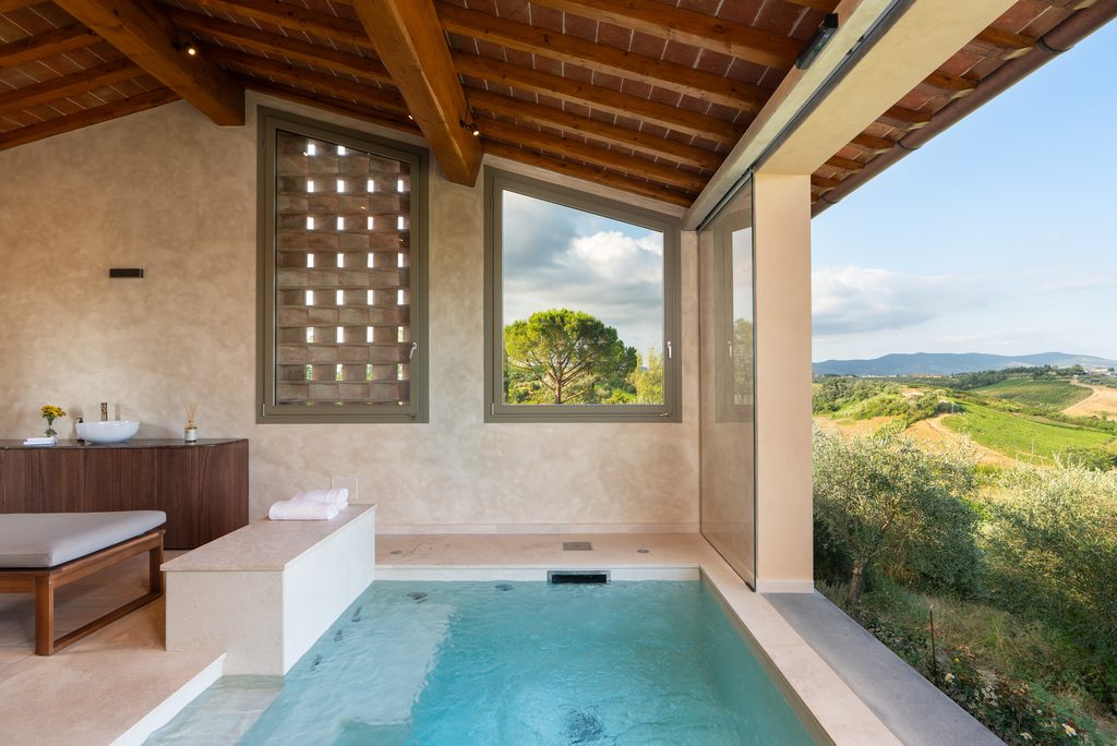 Vasca della piscina interna con bordo in pietra chiara, soffitto in travi di legno e cotto e vetrata aperta sulle vigne.