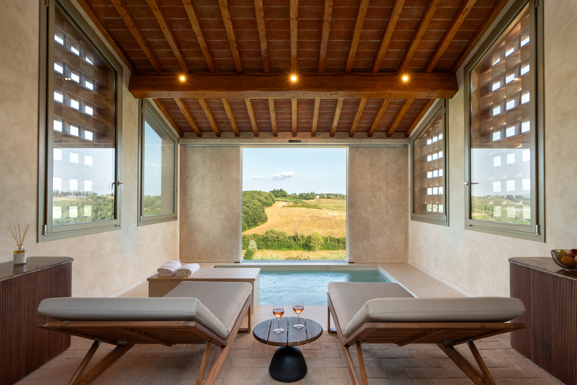 Piscina interna di Castelforno con due lettini, soffitto in travi di legno e cotto e grande apertura sulle colline di Cerreto Guidi.