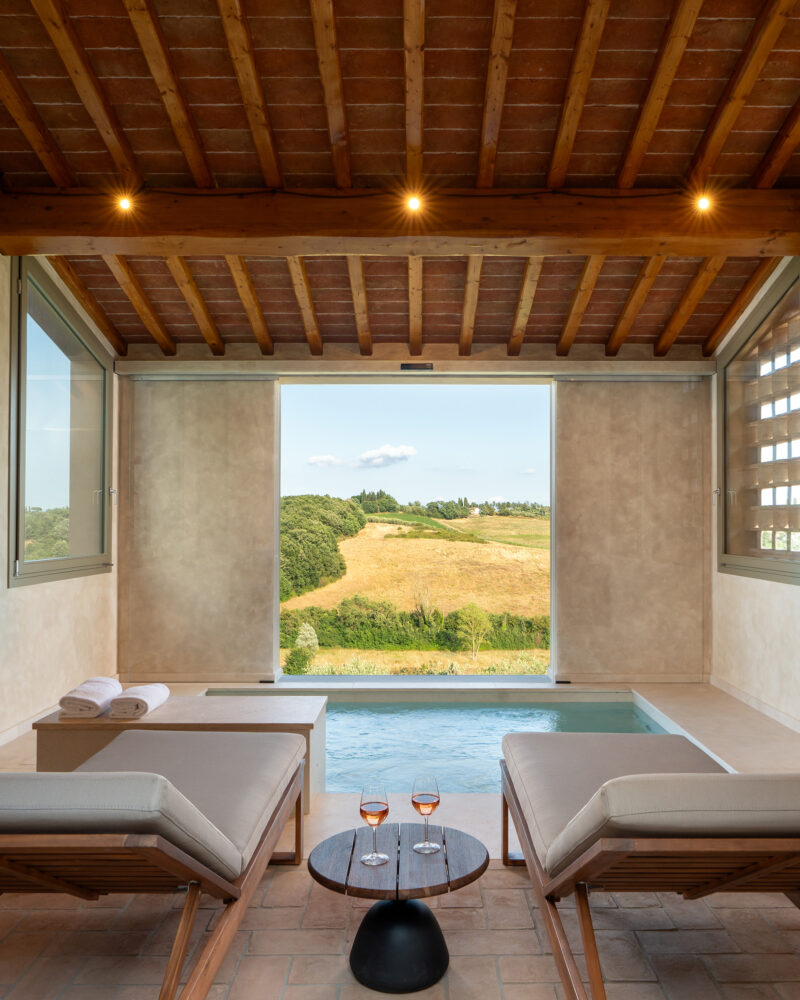 Piscina interna di Castelforno con due lettini, soffitto in travi di legno e cotto e grande apertura sulle colline di Cerreto Guidi.