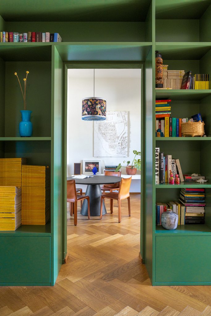 Libreria verde a tutta altezza attraversata dal passaggio verso la zona pranzo; in fondo il tavolo rotondo e la lampada a sospensione in tessuto.