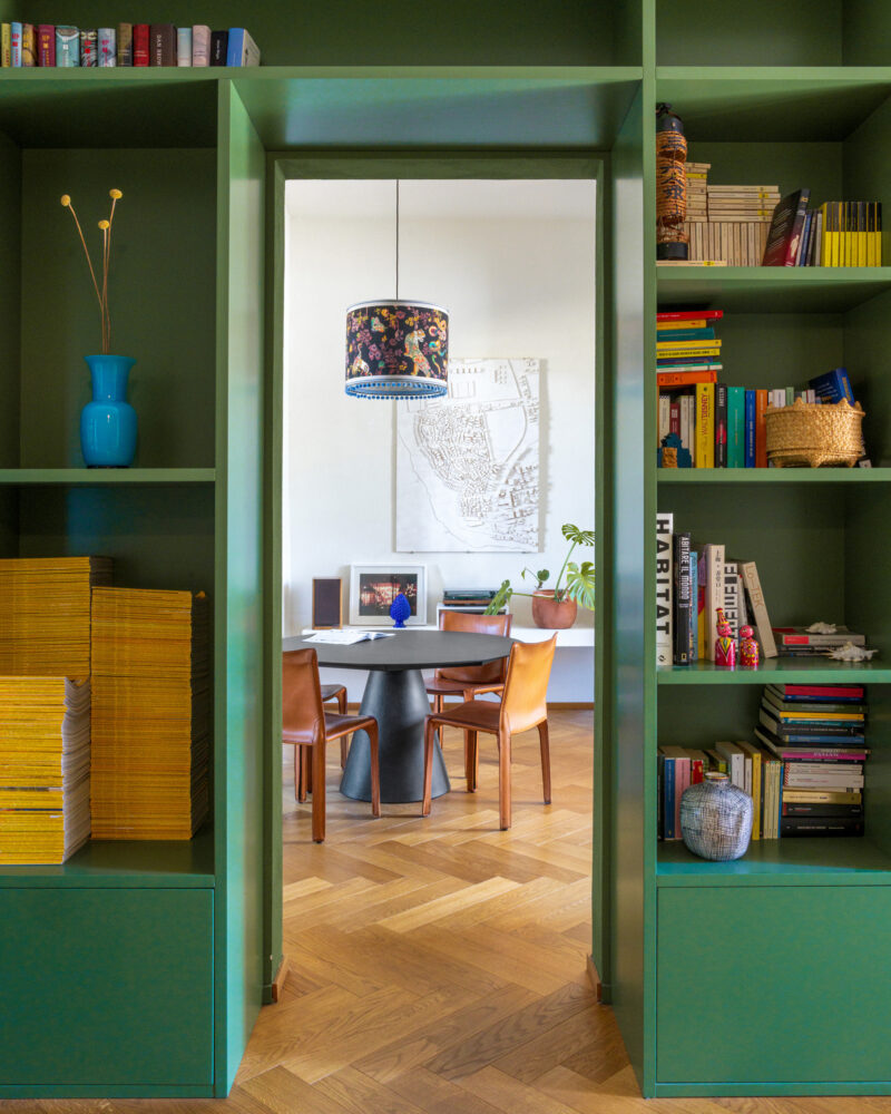 Libreria verde su misura che incornicia il passaggio verso la zona pranzo, con il tavolo rotondo nero e le sedie in cuoio.