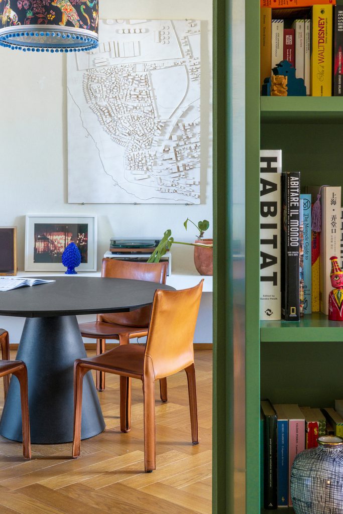 Zona pranzo con tavolo rotondo nero e sedie in cuoio accanto alla libreria verde; alla parete un pannello bianco in rilievo con la pianta di una città.