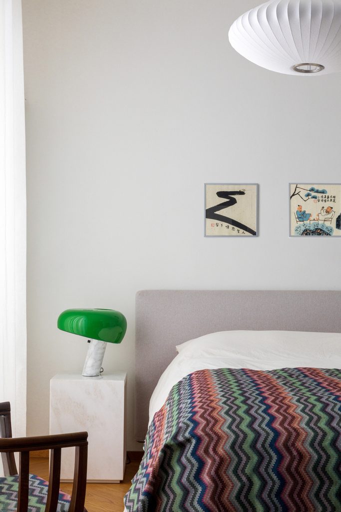 Camera da letto con testiera in tessuto, coperta a zigzag multicolore e lampada da tavolo verde su un comodino in pietra chiara.