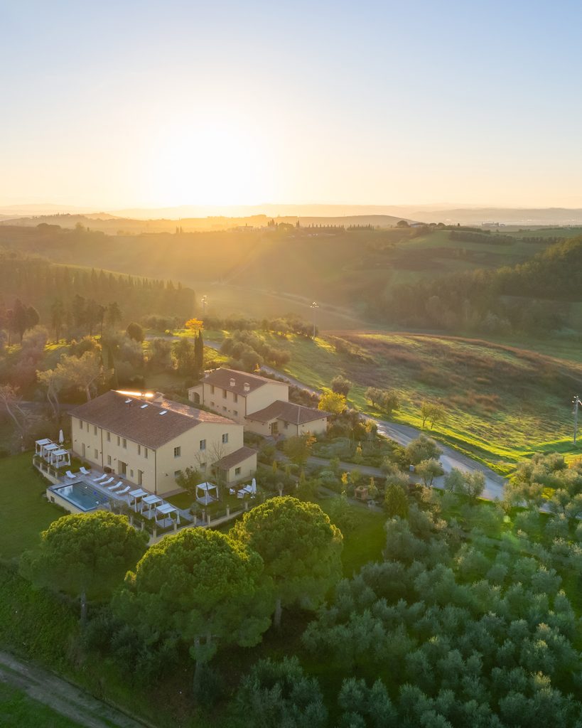 Vista aerea in controluce del casale di Podere San Martino con la piscina, le pergole e i pini domestici, tra oliveti e colline.