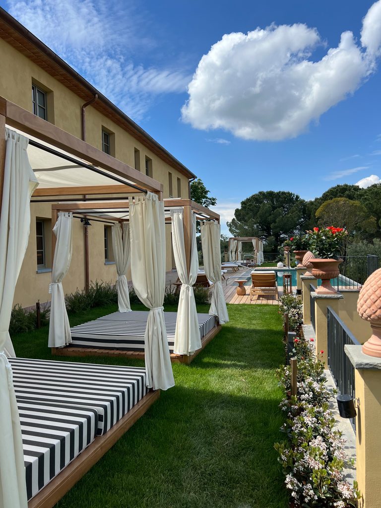 Pergole in legno con tende chiare e materassi a righe sul prato, lungo il casale; in fondo la pedana in legno e la piscina.