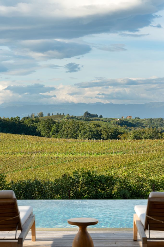 Vigne e colline viste dal bordo della piscina a sfioro, tra due lettini in legno.