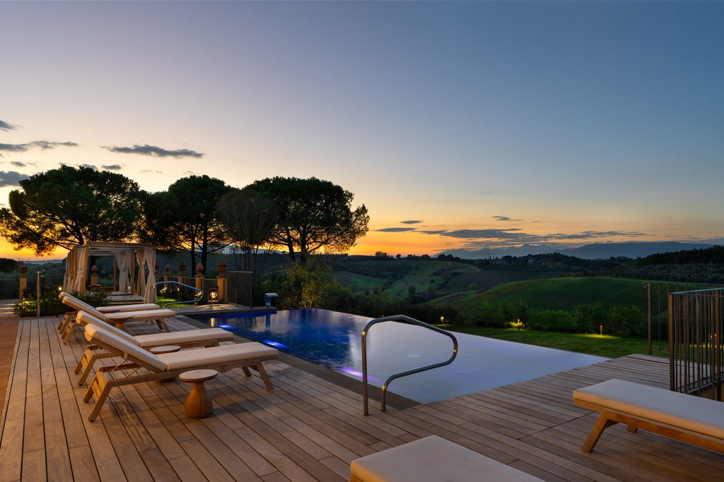 Piscina a sfioro illuminata al tramonto, con la pedana in legno, i lettini e una pergola sotto i pini.
