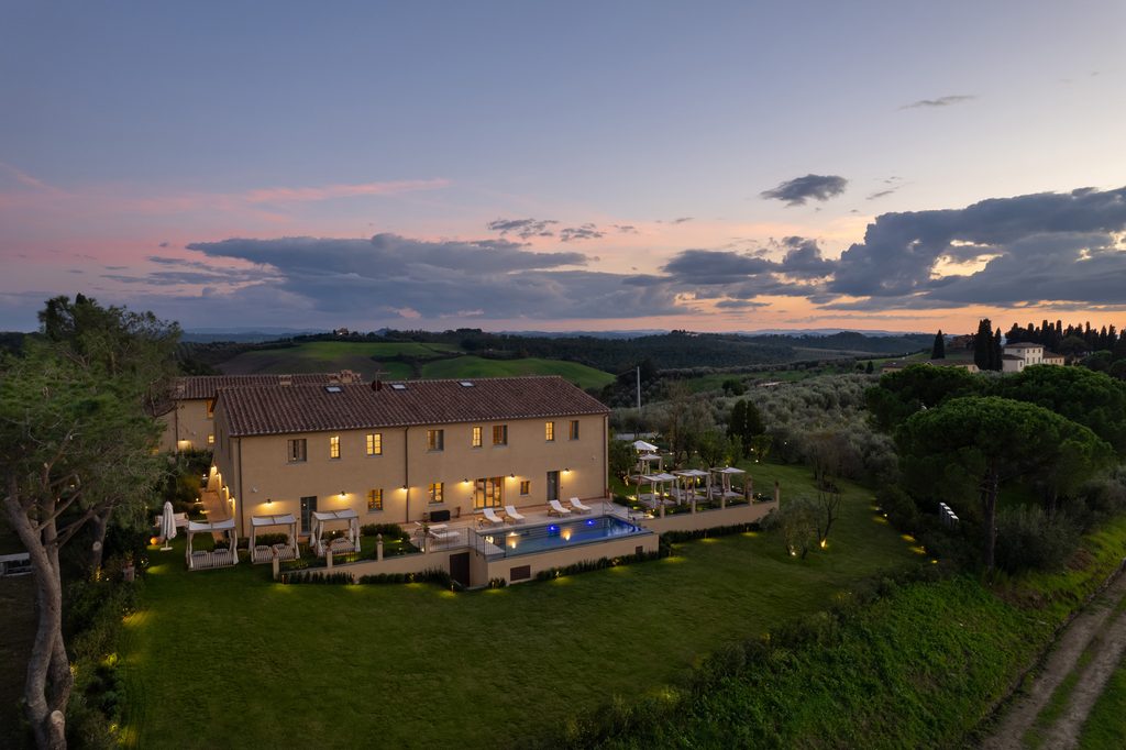 Vista aerea di Podere San Martino al crepuscolo, con il giardino e la piscina illuminati.